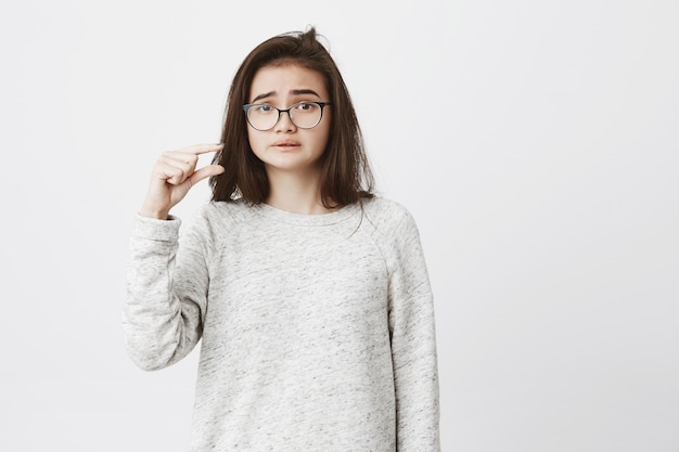Portrait de malheureuse femme mignonne avec des lunettes montrant quelque chose de petit ou petit