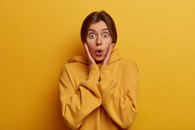 Le portrait d'une mannequin femme millénaire étonnée saisit le visage, regarde les yeux sortis, voit quelque chose de bouleversant, écoute une histoire choquante époustouflante, pose en jaune, habillé avec désinvolture. Oh mon Dieu vraiment