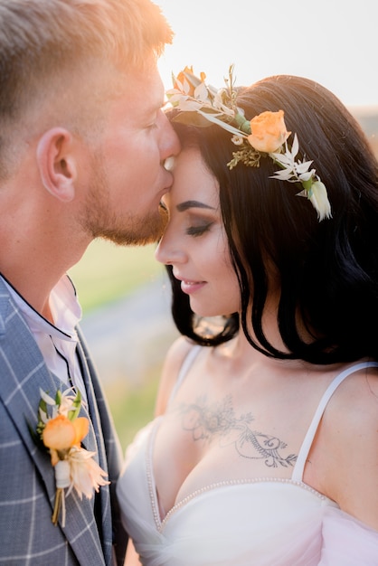 Portrait de marié qui embrasse la mariée tatouée avec décolleté ouvert et tendre couronne faite de fleurs fraîches