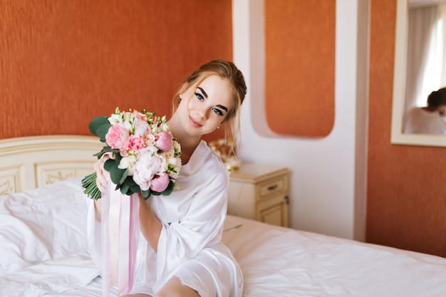 Portrait mariée assez heureuse en peignoir blanc avec des fleurs sur le lit le matin.