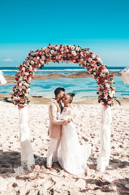 Photo gratuite portrait de la mariée et le marié posant près de l'arche tropicale de mariage sur la plage derrière le ciel bleu et la mer. couple de mariage