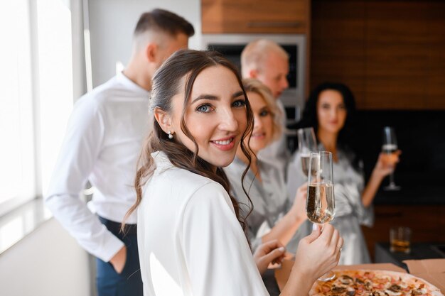 Portrait de la mariée souriante tenant un verre de champagne avec une coiffure de mariage regardant la caméra marié avec des invités flous sur fond Célébrer le matin des fiançailles dans l'appartement