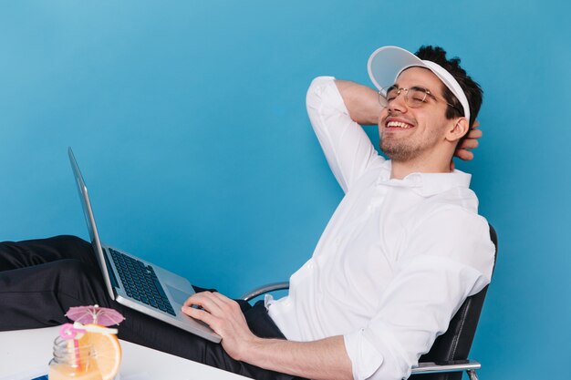 Portrait de mec joyeux à lunettes, casquette blanche et chemise souriant et tenant un ordinateur portable.