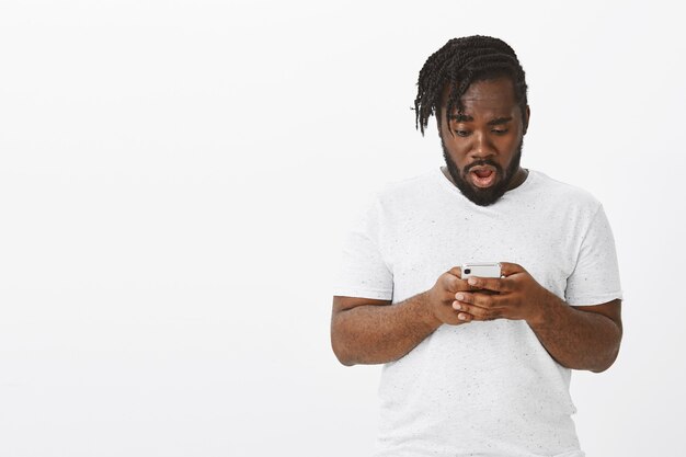 Portrait de mec surpris avec des tresses posant contre le mur blanc avec son téléphone