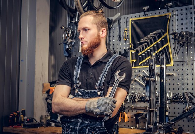 Portrait d'un mécanicien de vélo barbu aux bras croisés tenant une clé de tasse sur fond de support d'outil dans un atelier.