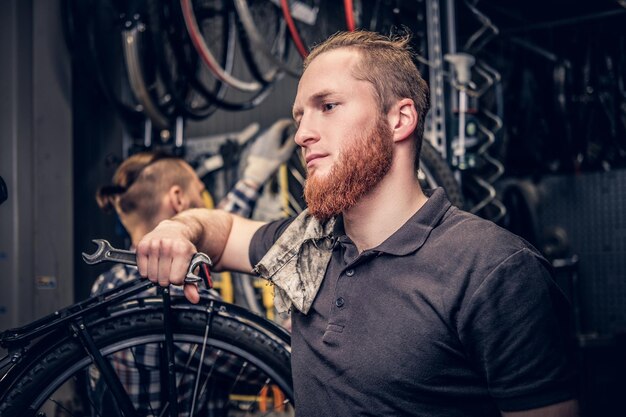 Portrait d'un mécanicien de vélo barbu à tête rouge dans un atelier avec des pièces de vélo et une roue sur fond.