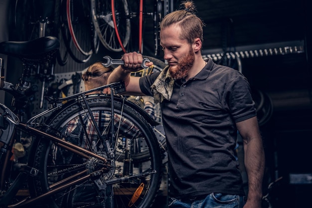 Portrait d'un mécanicien de vélo barbu à tête rouge dans un atelier avec des pièces de vélo et une roue sur fond.