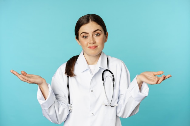 Portrait d'un médecin, femme stagiaire haussant les épaules, souriant et disant désolé, ne sais pas, étant désemparé, debout sur fond bleu.