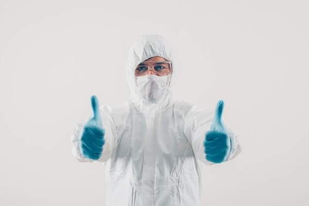 Portrait d'un médecin à fond clair montrant les pouces vers le haut dans des gants médicaux et une combinaison de protection