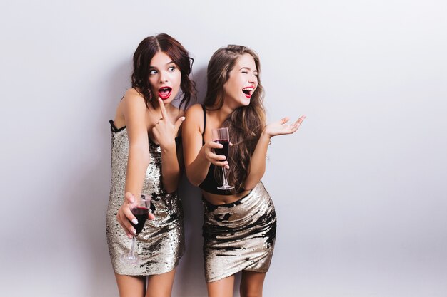 Portrait des meilleurs amis, jolies filles s'amusant, fête à l'intérieur et boire du vin rouge, fou, riant. Porter des robes brillantes à la mode, une jupe, une coiffure ondulée. Isolé.