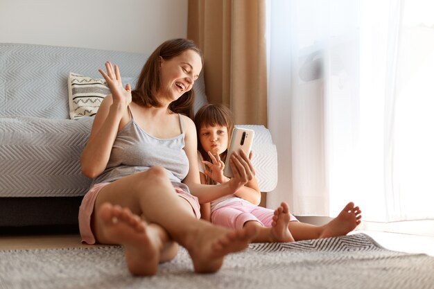 Portrait d'une mère blogueuse extrêmement heureuse diffusant en direct avec sa fille, agitant la main aux abonnés, exprimant des émotions positives, riant joyeusement.