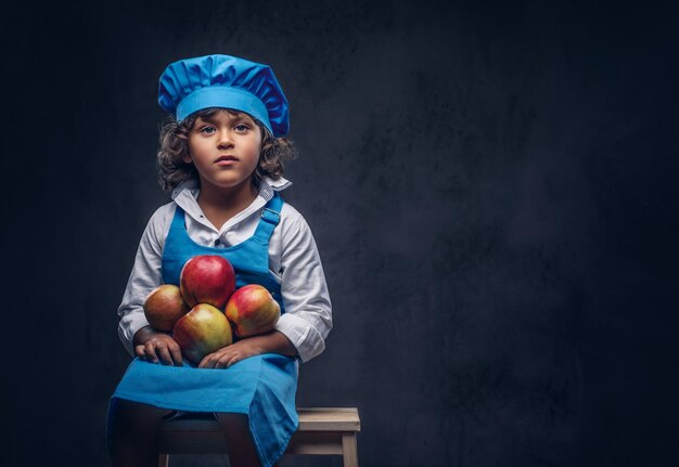 Portrait d'un mignon petit garçon aux cheveux bouclés bruns vêtu d'un uniforme de cuisinier bleu tient des pommes assis sur un tabouret en bois dans un studio. Isolé sur le fond texturé sombre.