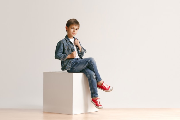 Le portrait de mignon petit garçon enfant dans des vêtements de jeans élégants regardant la caméra contre le mur blanc du studio. Concept de mode pour enfants