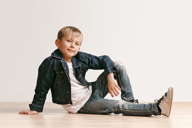 Le portrait de mignon petit garçon enfant dans des vêtements jeans élégants regardant la caméra contre le mur blanc du studio.