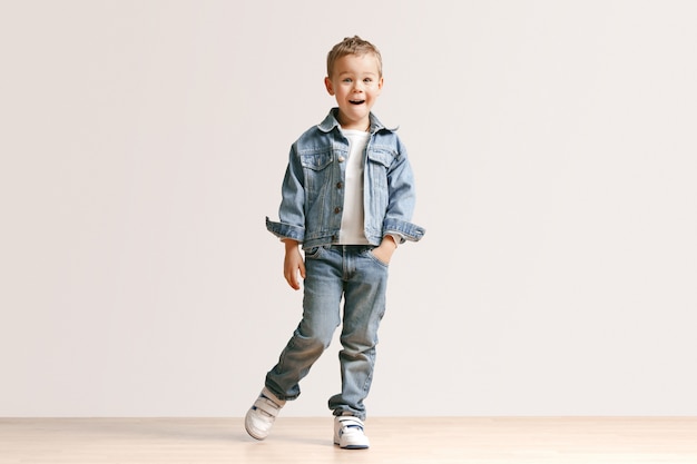 Le portrait de mignon petit garçon enfant dans des vêtements jeans élégants regardant la caméra contre le mur blanc du studio.