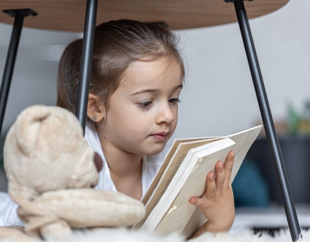 Portrait d'une mignonne petite fille lisant un livre à la maison, allongée sur le sol avec son jouet préféré.