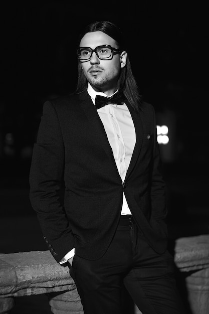 Portrait de mode élégant jeune homme aux cheveux longs. Modèle masculin attrayant et beau en costume noir avec moustache dans la rue la nuit