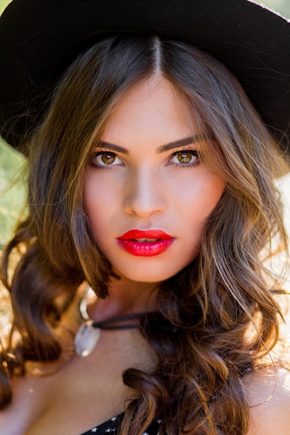 Portrait de mode d'été d'une femme élégante aux cheveux ondulés parfaits dans un chapeau noir élégant et élégant et un maquillage lumineux posant dans le parc. Style de rue.