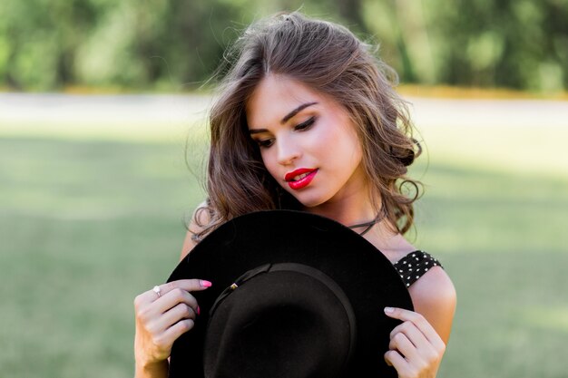 Portrait de mode d'été d'une femme élégante aux cheveux ondulés parfaits dans un chapeau noir élégant et élégant et un maquillage lumineux posant dans le parc.