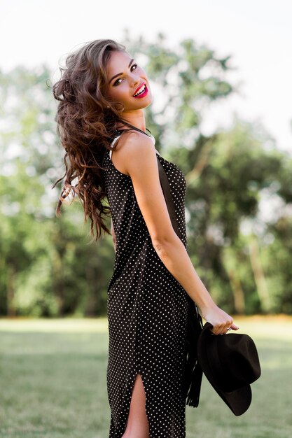 Portrait de mode d'été d'une femme élégante aux cheveux ondulés parfaits dans un chapeau noir élégant et élégant et un maquillage lumineux posant dans le parc.