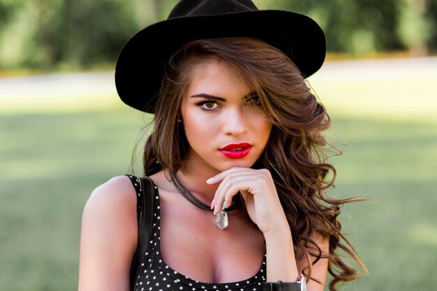 Portrait de mode d'été d'une femme élégante aux cheveux ondulés parfaits dans un chapeau noir élégant et élégant et un maquillage lumineux posant dans le parc.