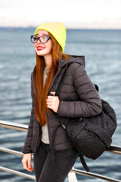 Portrait de mode hipster en plein air de jeune jolie femme aux longs poils roux, posant près du bord de mer, voyageant seul avec sac à dos, temps froid, look d'hiver de style rue élégant, chapeau, veste, pull.