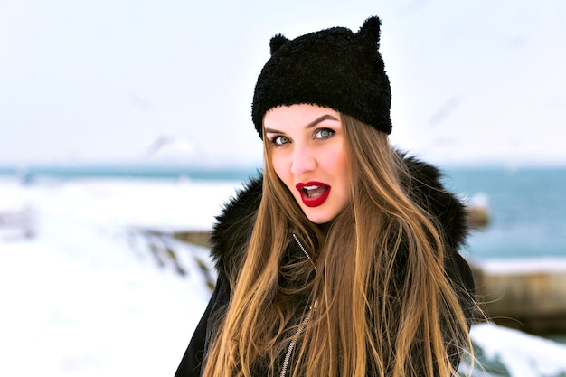 Photo gratuite portrait de mode d'hiver de femme blonde sensuelle, lèvres rouges pleines, beaucoup de neige, chapeau drôle, manteau élégant, expédition de voyage d'hiver, poils longs, temps venteux, mer de glace incroyable.