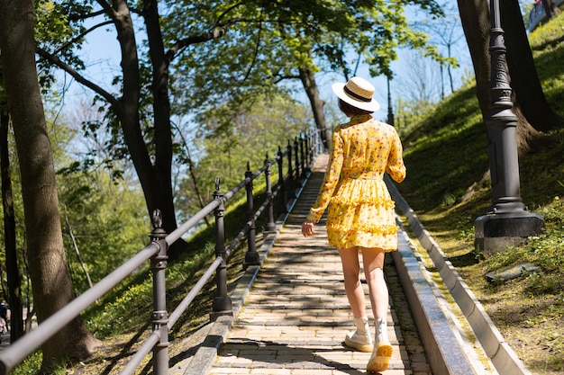 Portrait de mode en plein air de femme en robe d'été jaune et chapeau marchant sur le chemin dans le parc vue arrière