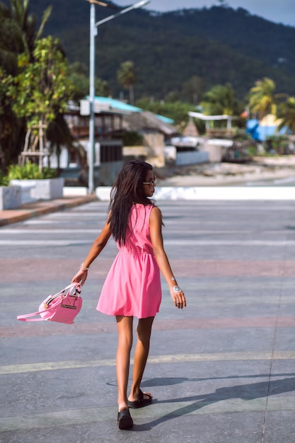 Portrait de mode en plein air d'une femme thaïlandaise asiatique assez mince posant dans la rue, portant une mini jolie robe rose, des sandales, des lunettes de soleil et un sac de couleur assortie, humeur de voyage.