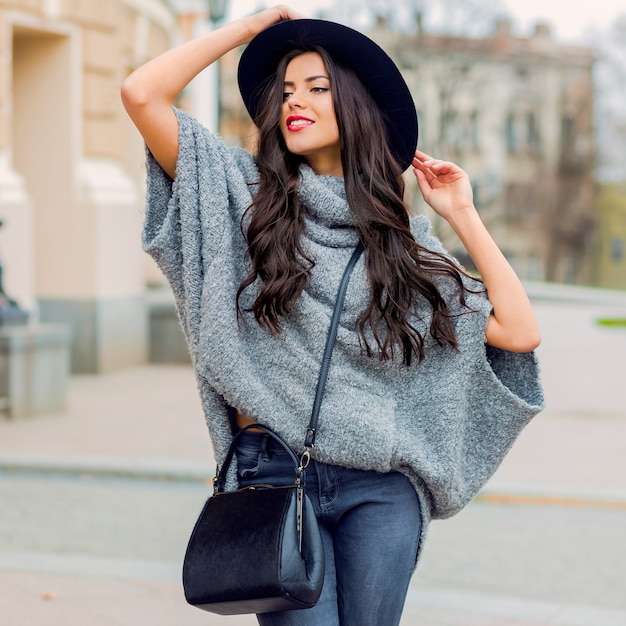 Portrait de mode en plein air de glamour sensuelle jeune femme élégante portant une tenue d'automne à la mode, un chapeau noir, un pull gris et un sac en cuir. Lèvres rouge vif. Contexte de la vieille ville.