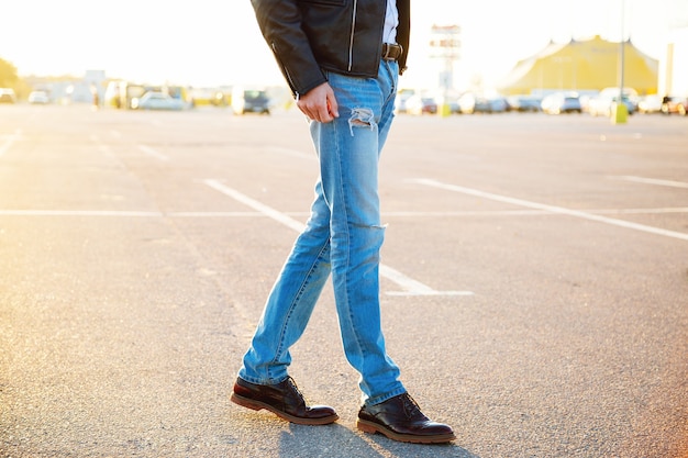 Portrait de mode en plein air urbain d'un jeune homme élégant hipster portant un pantalon en jean veste de motard en cuir et des chaussures vintage posant au soleil du soir de stationnement de campagne.