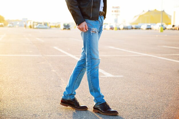 Portrait de mode en plein air urbain d'un jeune homme élégant hipster portant un pantalon en jean veste de motard en cuir et des chaussures vintage posant au soleil du soir de stationnement de campagne.