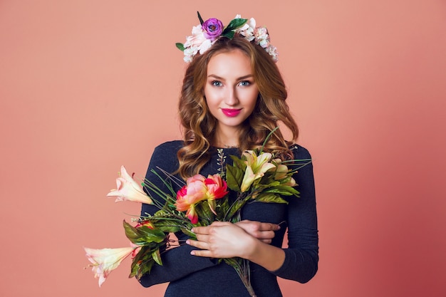 Portrait de mode romantique de printemps belle jeune femme aux longs cheveux blonds ondulés en guirlande de fleurs de printemps posant avec bouquet de fleurs sur fond rose.