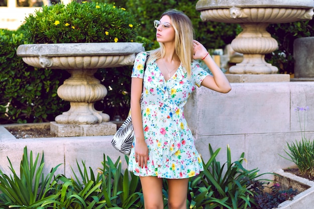 Portrait de mode de vie d'été de l'heureuse magnifique femme élégante blonde vêtue d'une élégante robe de thé floral tendance élégante et posant au vieux parc européen, ambiance de voyage.