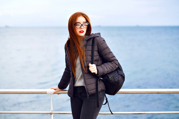 Portrait de mode de vie d'hiver de jolie femme au gingembre hipster marchant au bord de la mer, tenue élégante noire totale, temps froid et pluvieux, sac à dos, bottes en cuir, solitaire.