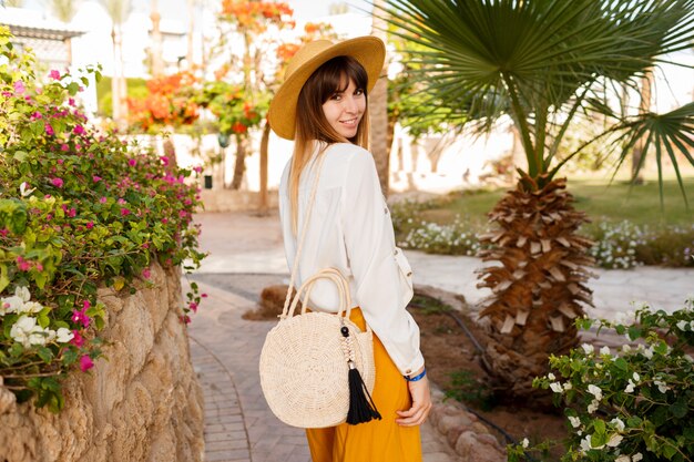 Portrait de mode de vie de jolie femme de race blanche en chapeau de paille, chemisier blanc et sac de style balinais marchant dans un jardin tropical.