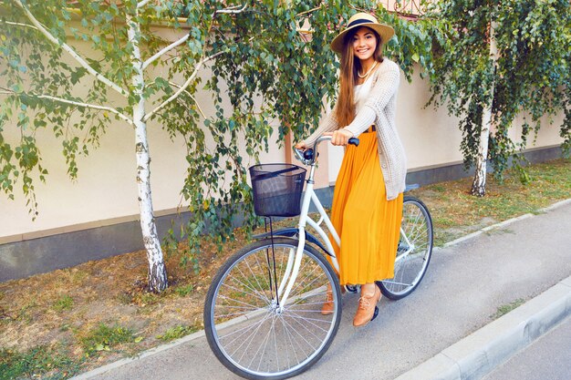 Portrait de mode de vie en plein air de la belle jeune fille brune chevauchant son vélo rétro sur la route avec des bouleaux. Porter un chapeau de vêtements élégant et un cardigan chaud. Humeur d'automne.