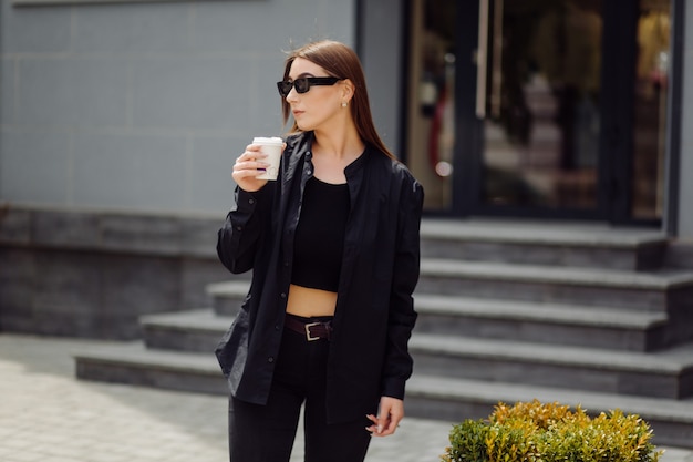 Portrait de mode de vie en plein air d'une superbe fille brune. Boire du café et marcher dans la rue de la ville.