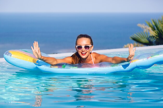 Photo gratuite portrait de mode de vie en plein air d'une superbe jeune femme s'amusant à la piscine à débordement avec vue imprenable sur l'île tropicale, portant un bikini lumineux et des lunettes de soleil, nageant sur un matelas pneumatique.
