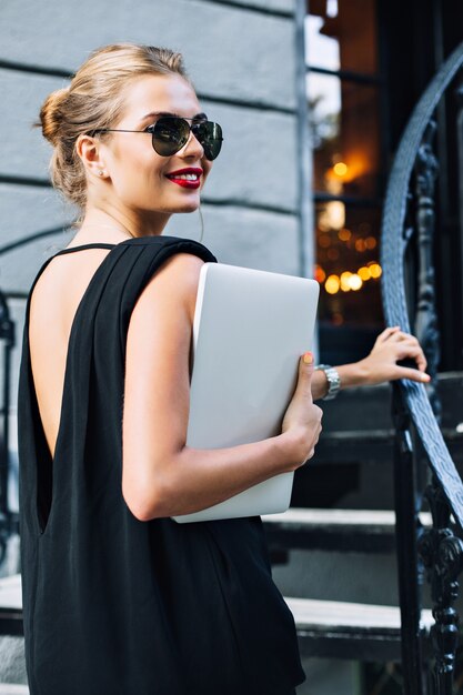 Portrait modèle attrayant en robe noire avec dos nu dans les escaliers en plein air. Elle sourit à côté.