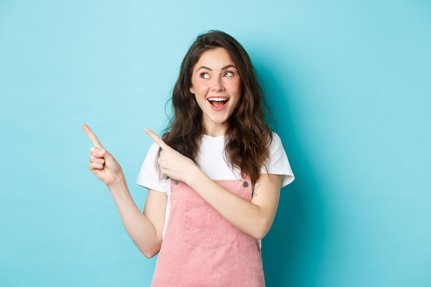 Portrait d'un modèle féminin souriant et heureux avec un blush glamour, pointant du doigt vers la gauche et regardant l'offre promotionnelle, montrant un espace de copie publicitaire, fond bleu