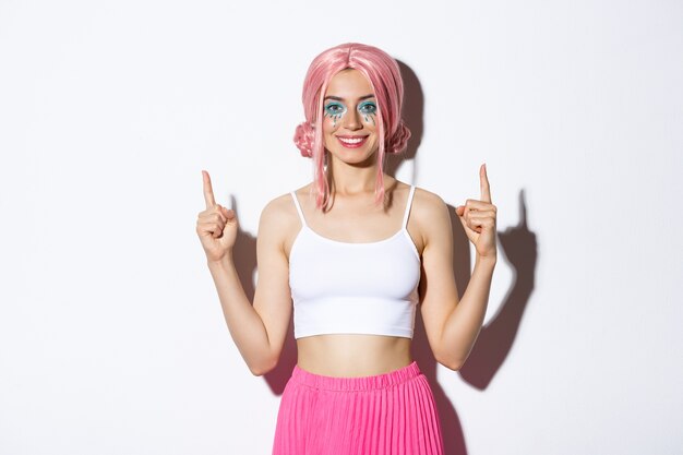 Portrait de modèle féminin souriant heureux avec perruque de fête et maquillage halloween glamour, pointant les doigts vers le haut, montrant votre logo, debout.