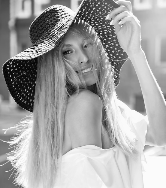 Portrait de modèle femme blonde sensuelle vêtue d'une robe blanche et d'un chapeau de plage d'été posant sur le fond de la rue derrière le coucher du soleil