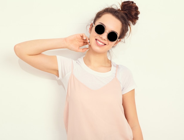 Portrait de modèle de jeune fille belle jeune femme brune avec du maquillage nude dans des vêtements colorés hipster rose d'été en lunettes de soleil posant près du mur. regardant joyeusement