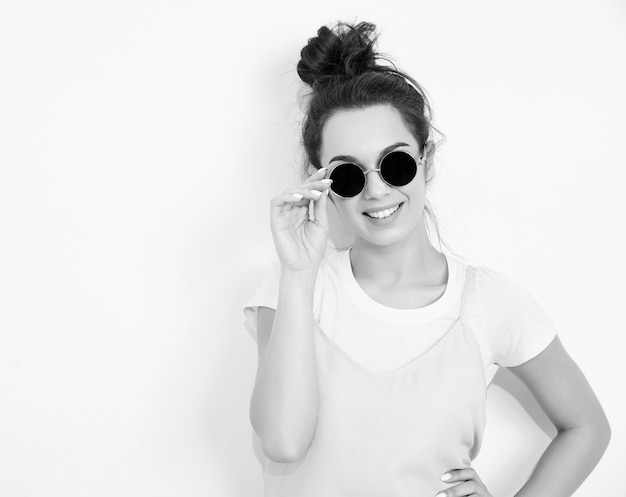 Portrait de modèle de jeune fille belle jeune femme brune avec du maquillage nude dans des vêtements colorés hipster rose d'été en lunettes de soleil posant près du mur. regardant joyeusement