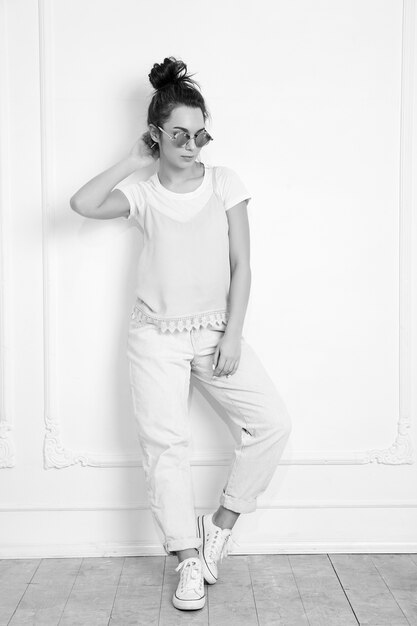 Portrait de modèle de jeune fille belle jeune femme brune avec du maquillage nude dans des vêtements colorés hipster rose d'été en lunettes de soleil posant près du mur. Toute la longueur