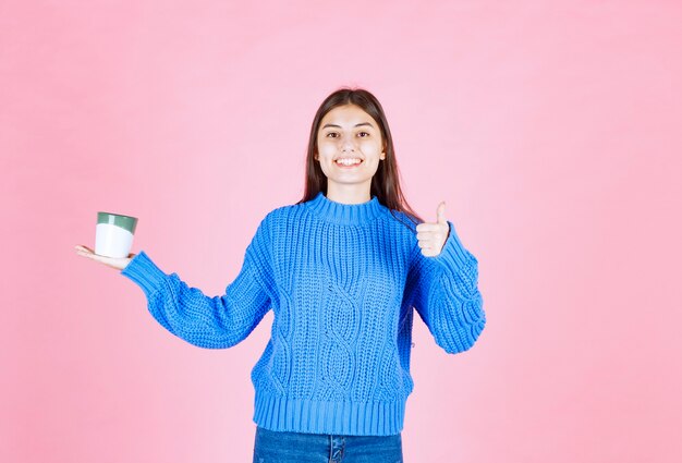 Portrait d'un modèle de jeune fille avec une tasse montrant un pouce vers le haut sur un mur rose.