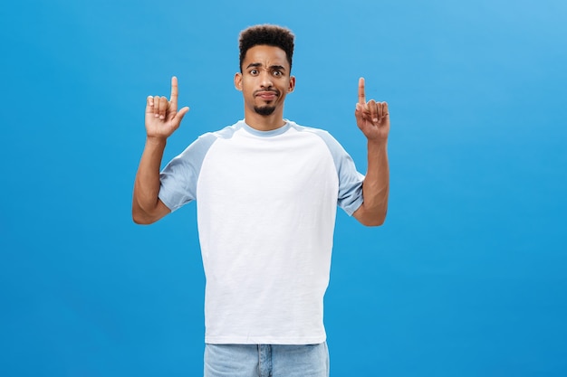 Portrait d'un modèle masculin afro-américain intense et douteux mécontent en t-shirt décontracté souriant et fronçant les sourcils de déception et de doute pointant vers quelque chose de suspect sur le mur bleu