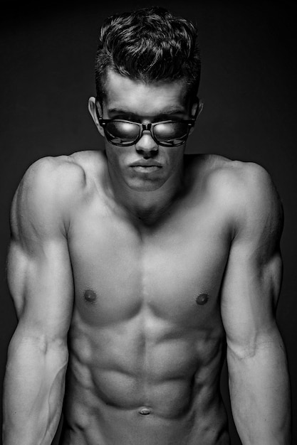 Photo gratuite portrait d'un modèle masculin génial dans des lunettes de soleil avec un corps musclé nu. isolé sur fond gris foncé.