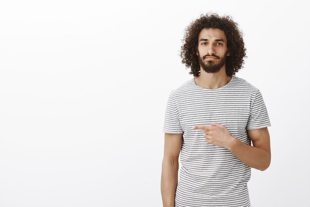 Portrait de modèle masculin oriental attrayant calme avec une coiffure frisée en t-shirt à la mode, pointant vers la gauche et souriant avec une expression décontractée